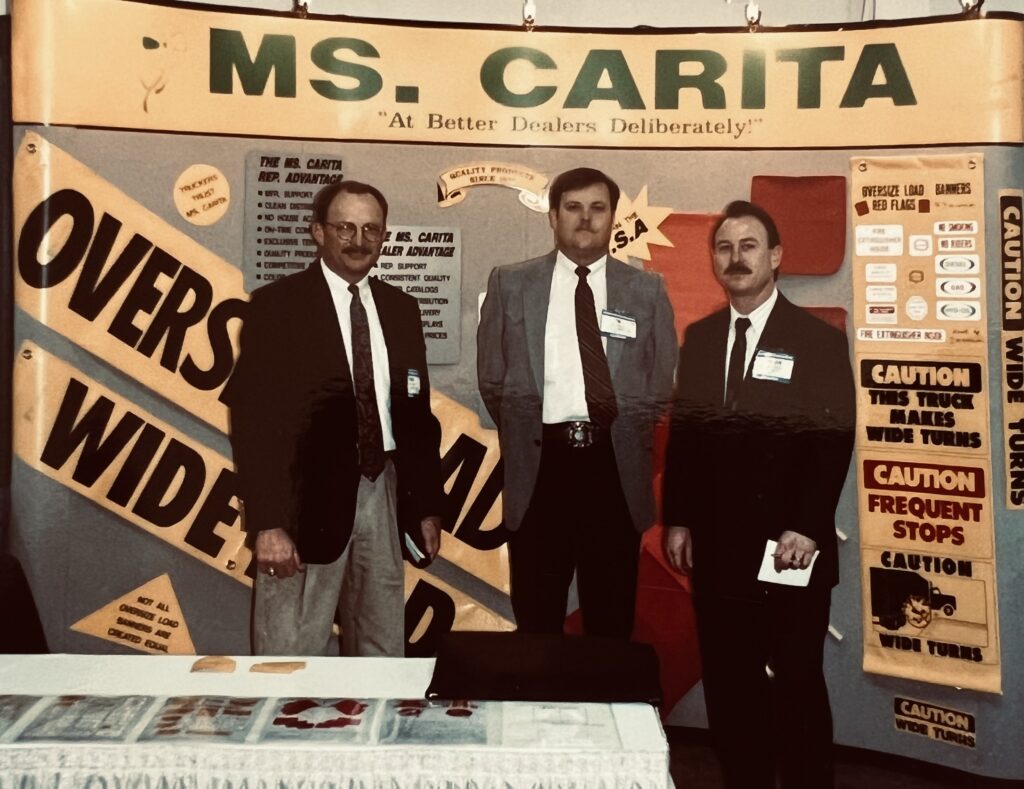 Gordon at a truck show in front of truck safety signs and flags at a tradeshow with two reps